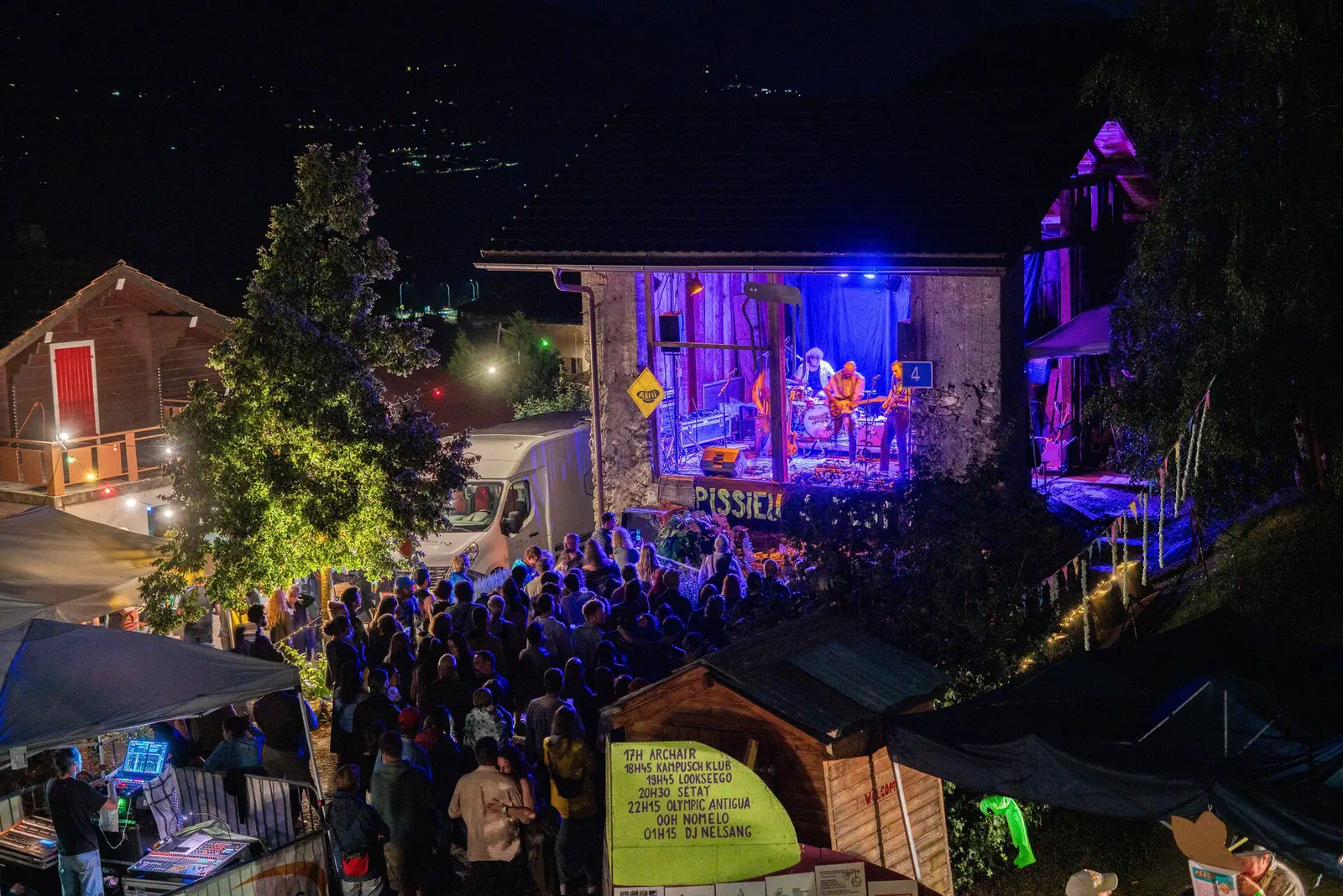Vue de nuit du Pissieu Festival 2025 : la grange du Musée valaisan des Bisses transformée en scène, éclairée en violet et bleu, un groupe joue, foule de spectateurs devant la scène, banderole &ldquo;PISSIEU&rdquo;.